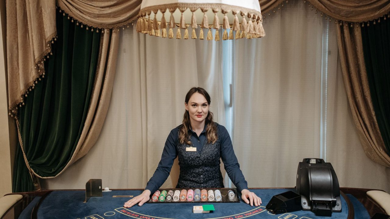 A smiling female casino dealer at a gaming table surrounded by chips and cards indoors.